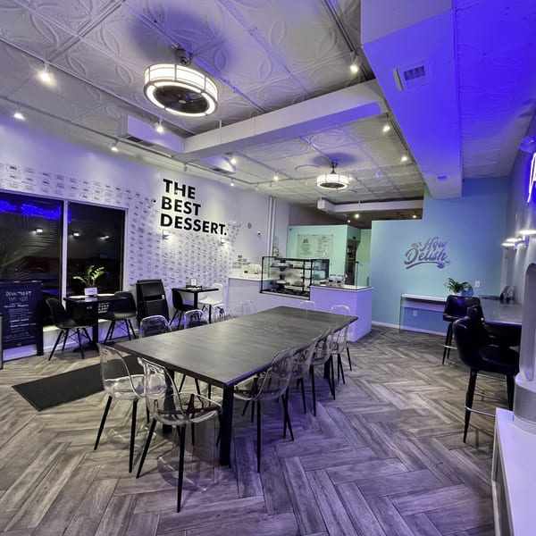 Interior of How Delish HD restaurant with tables, chairs, dessert display, and "The Best Dessert" wall art.