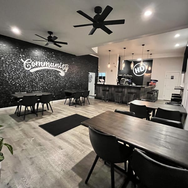 Interior of How Delish HD restaurant with dark tables, chairs, a bar, and a "Community" wall graphic.
