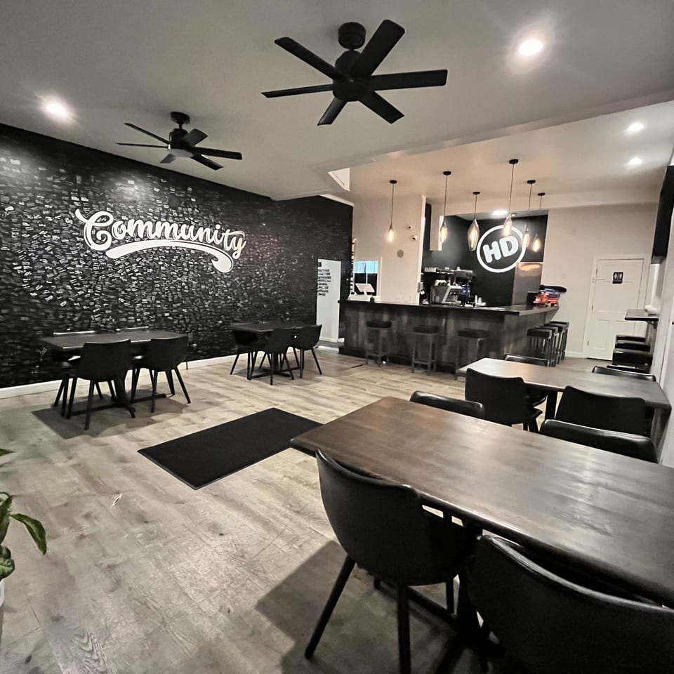 Interior of How Delish HD restaurant with dark tables, chairs, a bar, and a "Community" wall graphic.