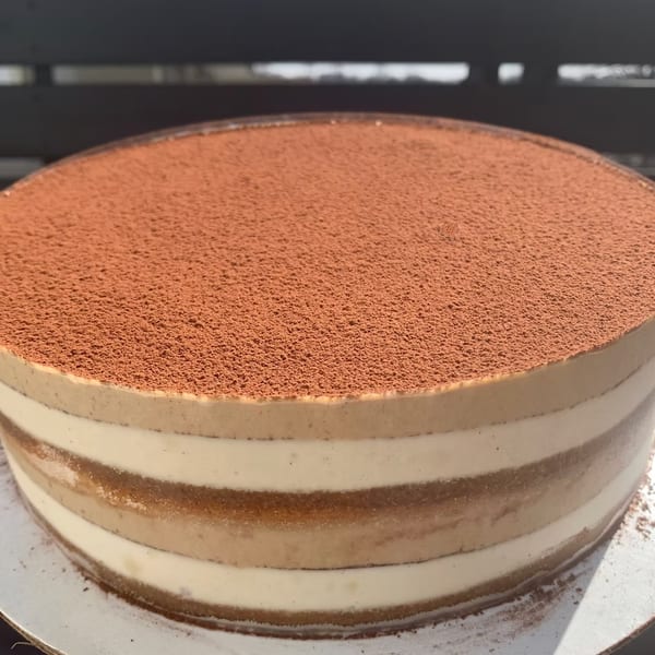 A round tiramisu cake with layers of cream and coffee-soaked sponge, topped with cocoa powder.