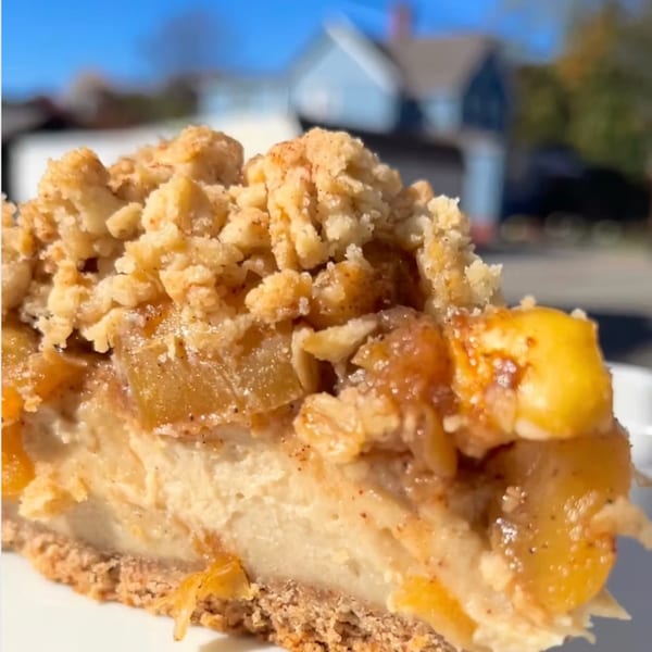 Close-up of a slice of apple crumble cheesecake with a house in the blurred background.
