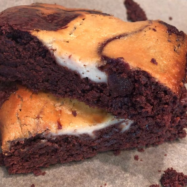 Two stacked cheesecake brownies with rich chocolate and creamy swirled topping on parchment paper.