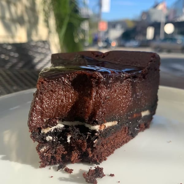 A slice of rich chocolate cake with frosting and a creamy white layer, on a white plate outdoors.