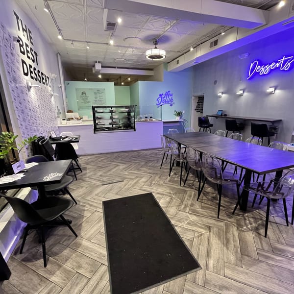 Interior of How Delish HD dessert shop with tables, chairs, a dessert display, and neon signs.