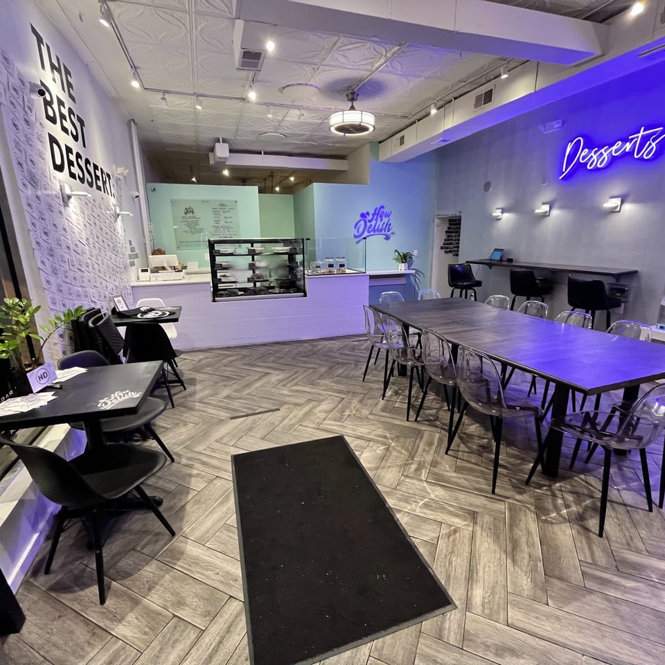 Interior of How Delish HD dessert shop with tables, chairs, a dessert display, and neon signs.