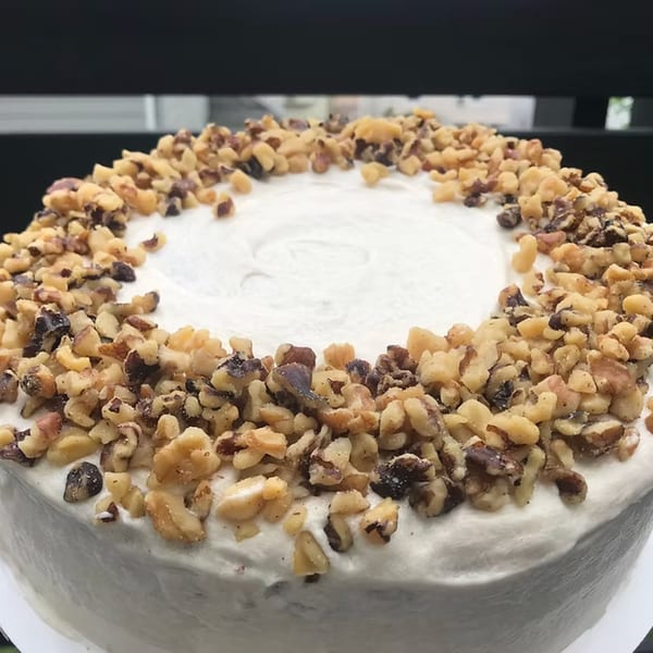 A round cake with white frosting, generously topped with chopped walnuts, from How Delish HD.