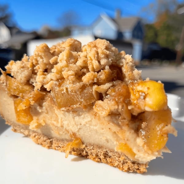 A slice of cheesecake with a crumbly topping and fruit chunks, on a white plate outdoors.