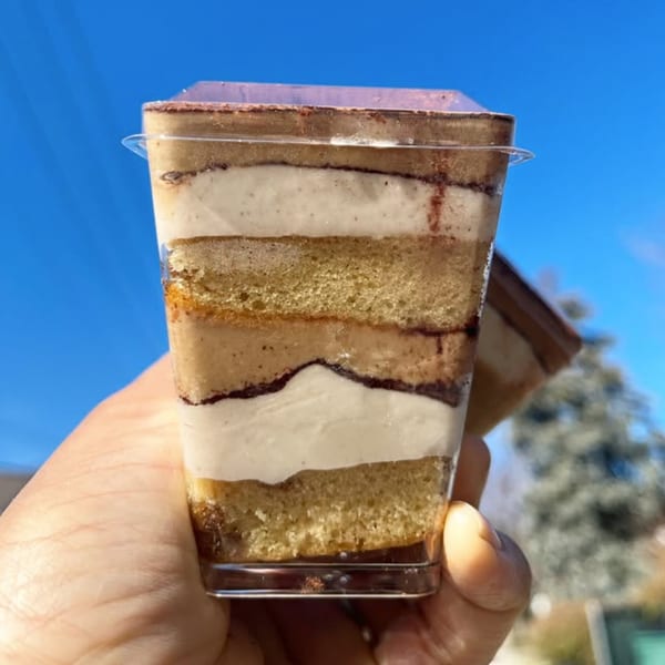 Hand holding a clear cup of layered tiramisu dessert with cake and cream, against a blue sky.