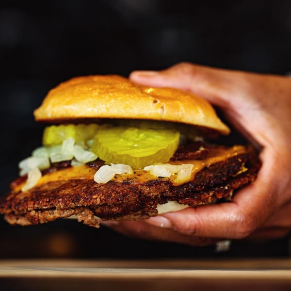 A hand holds a How Delish HD burger with a thick patty, cheese, pickles, and onions.