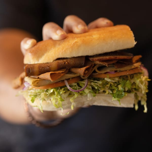 A hand holds a How Delish HD sandwich filled with meat, lettuce, tomatoes, and onions.
