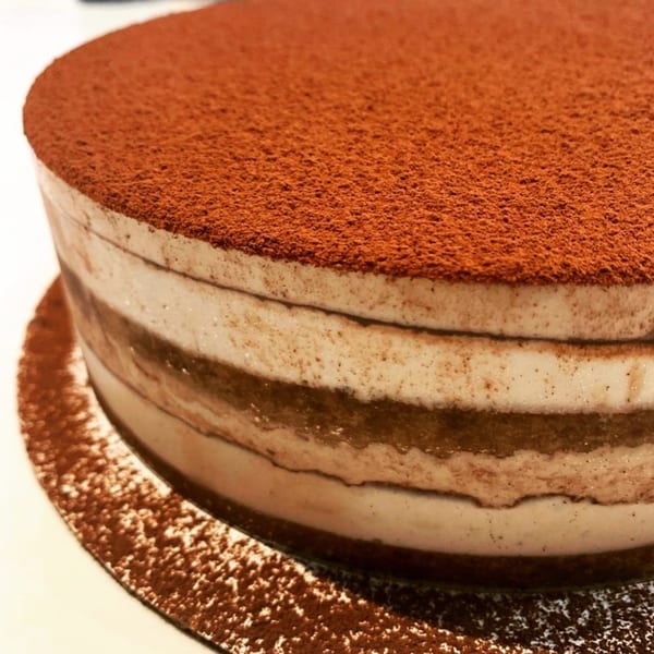 Close-up of a round tiramisu cake with layers of cream and coffee-soaked cake, topped with cocoa powder.