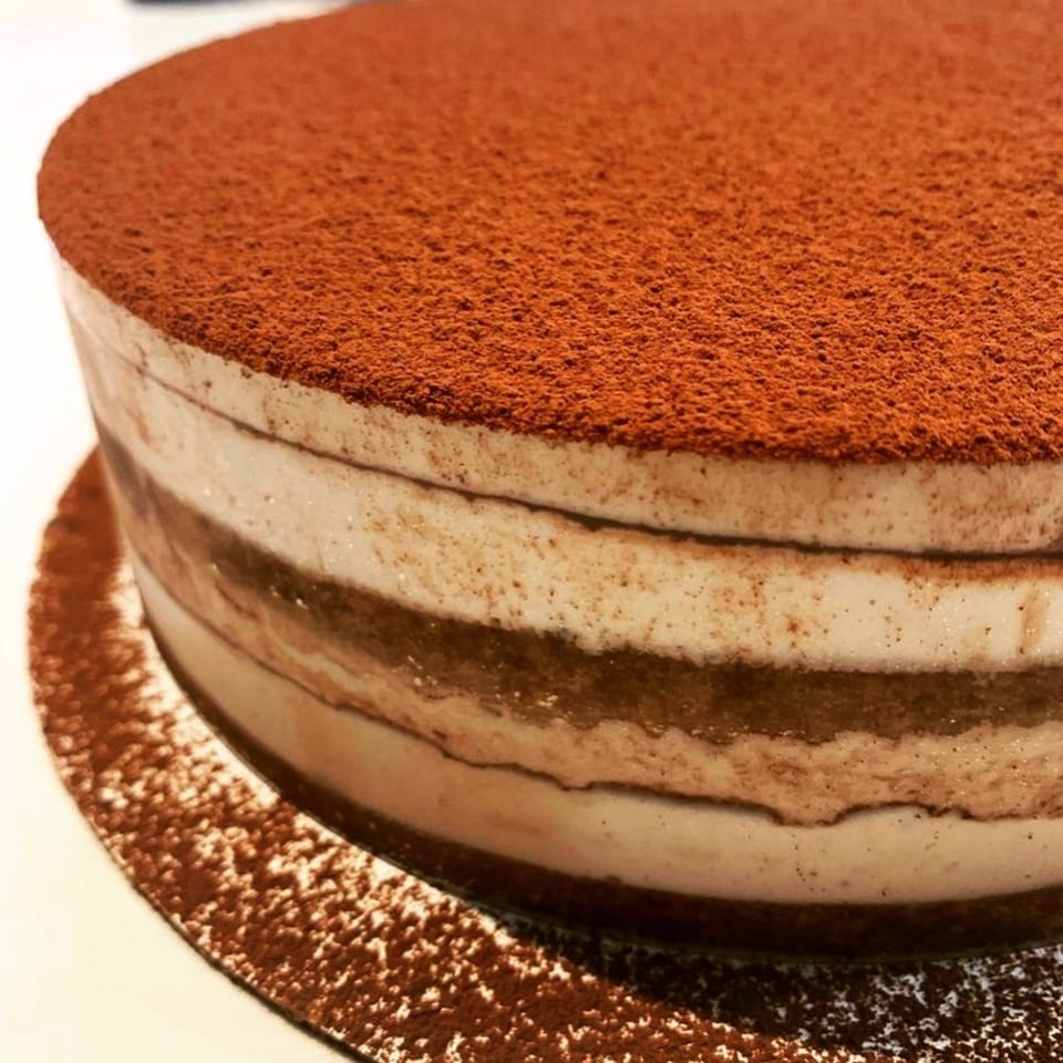 Close-up of a round tiramisu cake with layers of cream and coffee-soaked cake, topped with cocoa powder.