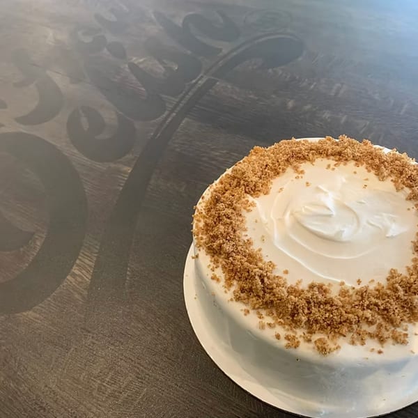 A round cake with white frosting and a border of brown crumble sits on a dark wooden table.