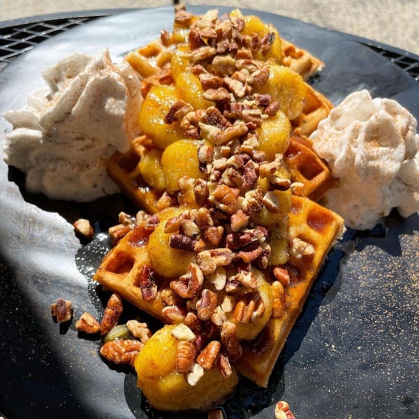 Golden waffles topped with caramelized bananas, pecans, and whipped cream on a dark plate.