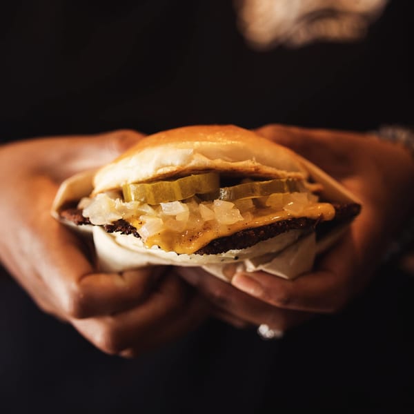 Hands holding a How Delish HD burger with pickles, onions, and cheese on a dark background.