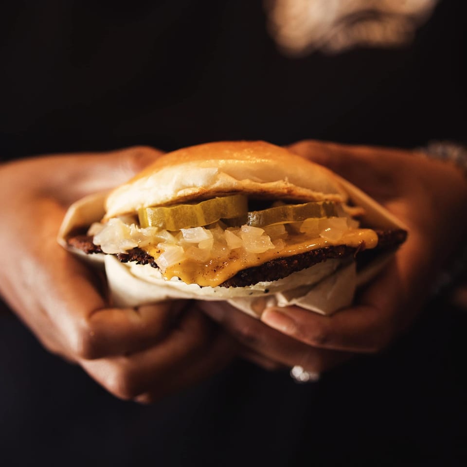 Hands holding a How Delish HD burger with pickles, onions, and cheese on a dark background.